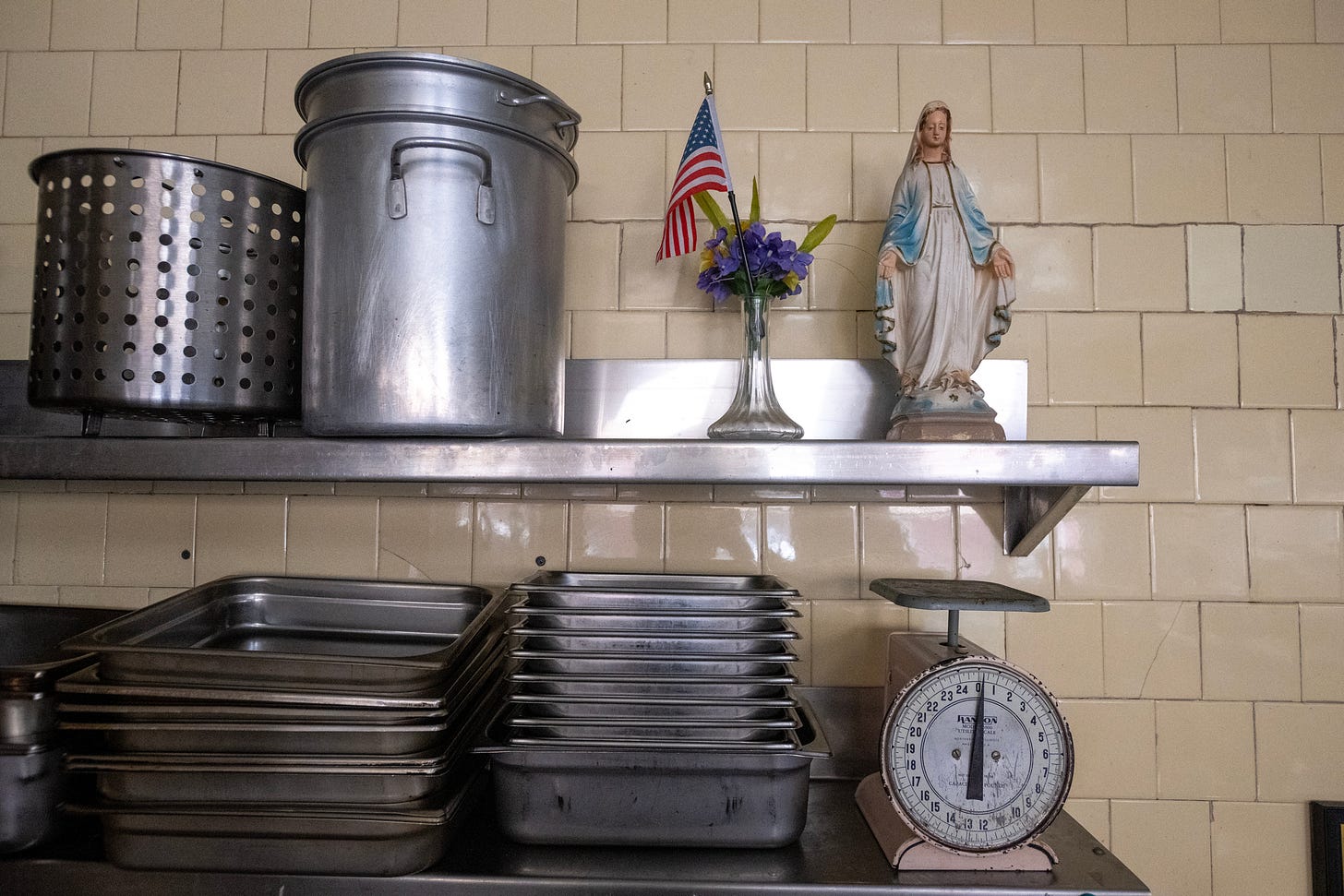 Old kitchen vessels, a scale, flowers, and a statue of Mary share a steel shelf