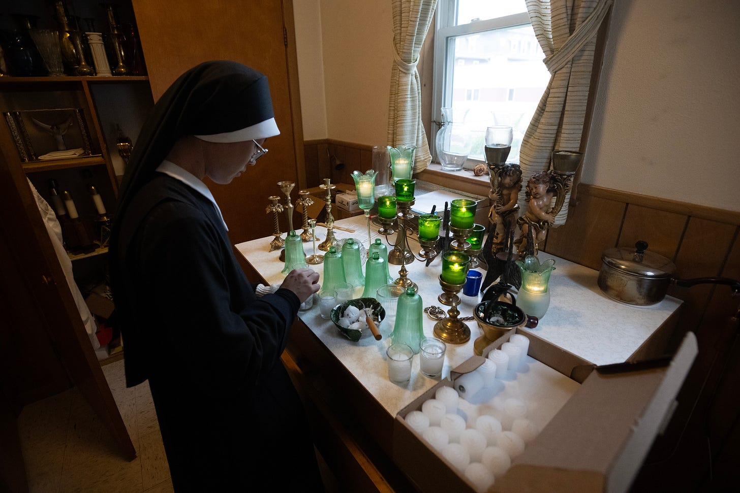 A Sister prepares candles and devotional vessels beside a window