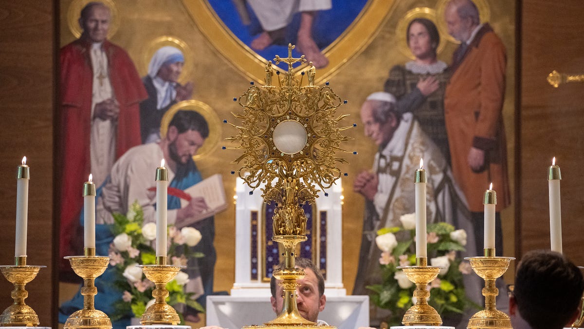 A gold monstrance stands between candles before an image of saints